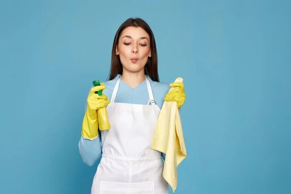 caucasian woman in rubber gloves and cleaner apron with cleaning rag and detergent sprayer on blue background.