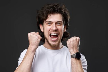 very excited young bearded man showing victory gesture holding fists raising hands up on gray background