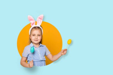 Sarı ve mavi arka planda renkli yumurtaları olan, Paskalya tavşanı kulaklı, gülümseyen küçük bir kız çocuğu. Mutlu Paskalyalar. Boşluğu kopyala