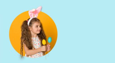 Gülümseyen kıvırcık saçlı, paskalya tavşanı kulaklı, sarı ve mavi arka planda renkli yumurtaları olan küçük kız çocuğu. Boşluğu kopyala