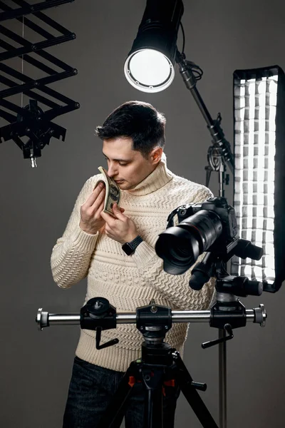 Professional handsome photographer with digital camera on tripod taking a lot of money and sniffing money on gray background with lighting equipment
