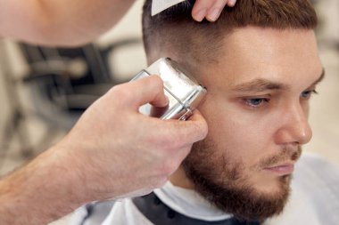 Berber dükkanındaki sakallı yakışıklının saçını kırpıcı ile kesiyor..