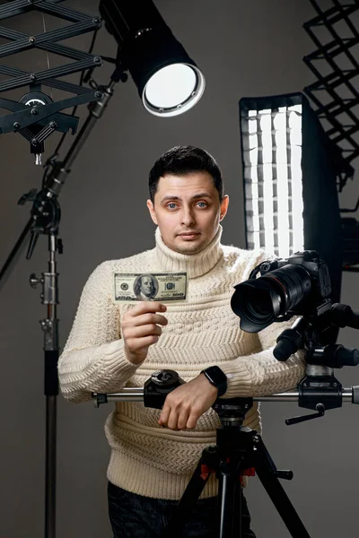 Professional handsome photographer with digital camera on tripod showing hundred dollar bill on gray background with lighting equipment