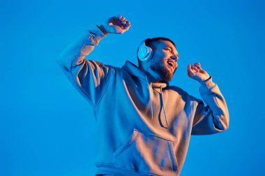 handsome bearded man with headphones in sweatshirt enjoying favorite music on blue neon background. Neon lighting