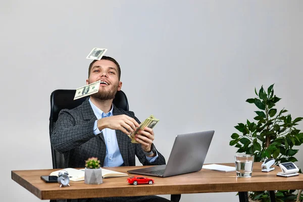 若い髭のビジネスマンがノートパソコンで働いてお金を投げながら机の上で椅子に座ってる 銀行券は空を飛ぶ — ストック写真