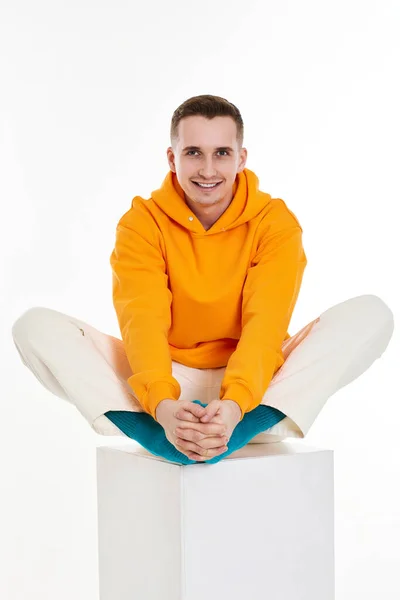 白い背景に白い立方体の上に座っているオレンジのスウェットシャツの青年 — ストック写真