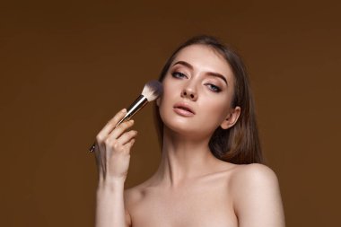 Bej arka planda makyaj fırçasıyla yanaklarına allık süren genç bir kadın. Boşluğu kopyala