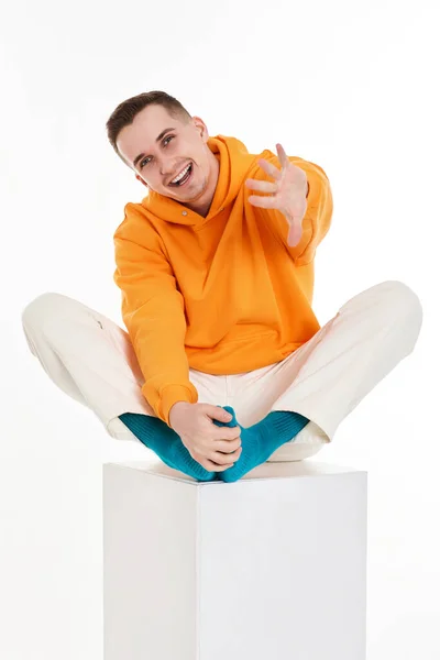 白い背景の白い立方体の上に座っているオレンジのスウェットシャツの若者 — ストック写真