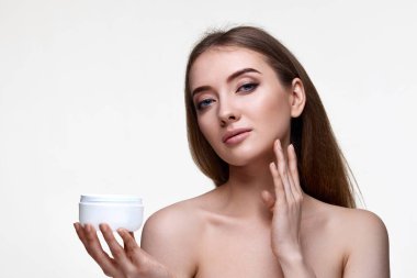 Güzel, gülümseyen genç bir kadın beyaz arka planda yüzüne krem sürüyor.