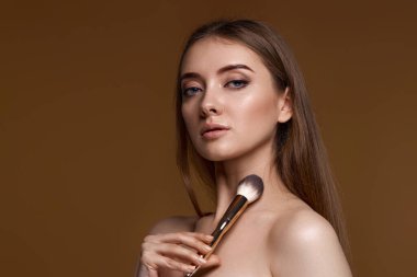 Bej arka planda makyaj fırçasıyla yanaklarına allık süren genç bir kadın. Boşluğu kopyala