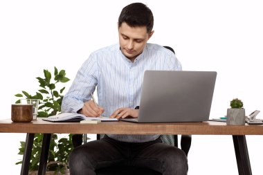 Mavi gömlekli iş adamı not alıyor, beyaz arka planda dizüstü bilgisayar kullanıyor.