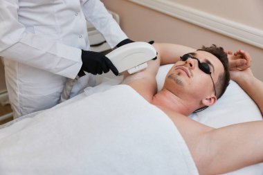 Genç adam güzellik merkezinde epilasyon alıyor. Profesyonel güzellik uzmanı erkek yüzünde lazer tüy çıkarma prosedürü uyguluyor..