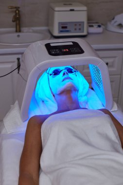 Koruyucu gözlüklü bir kadın terapiyle yüz bakımı yaptırıyor. Güzellik kliniğinde LED maske