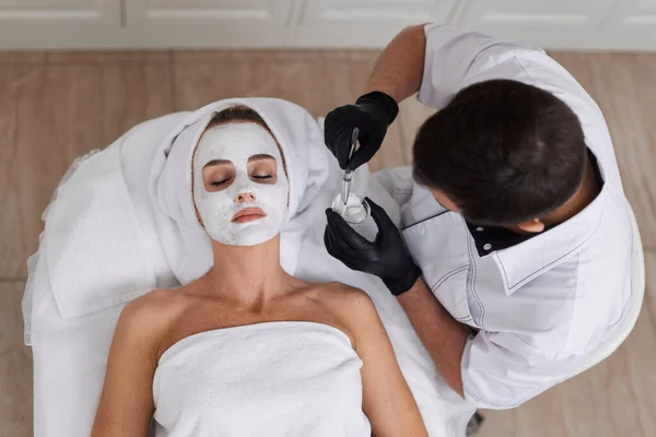 男美容师把面具贴在温泉沙龙美女的脸上 面部皮肤护理概念 — 图库照片