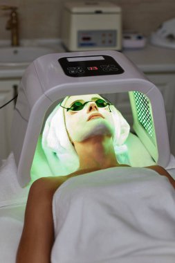 Koruyucu gözlüklü bir kadın terapiyle yüz bakımı yaptırıyor. Güzellik kliniğinde LED maske