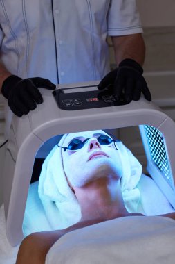 Koruyucu gözlüklü bir kadın terapiyle yüz bakımı yaptırıyor. Güzellik kliniğinde LED maske