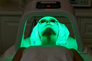 Koruyucu gözlüklü bir kadın terapiyle yüz bakımı yaptırıyor. Güzellik kliniğinde LED maske