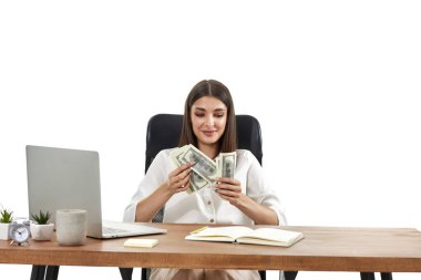 Mutlu iş kadını dizüstü bilgisayarda çalışıyor ve masa başında otururken elinde para tutuyor..