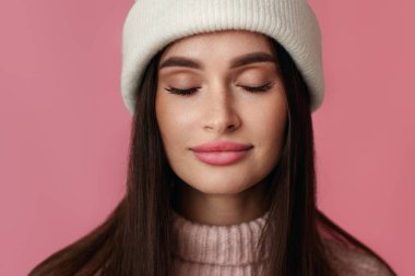 Beyaz şapkalı genç bir kadın portresi, pembe süveter, pembe arka planda gözlerini kaybediyor..