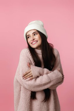 Beyaz şapkalı güzel bir kadın, kendine sarılıyor ve gülümsüyor. Pastel pembe arka planda mutlu hissediyor..