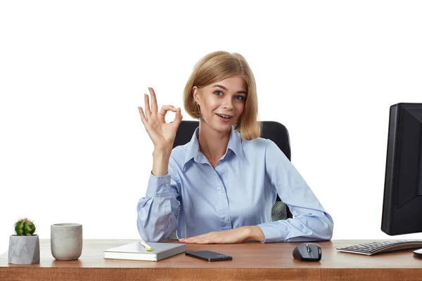 快乐的金发碧眼的女经理坐在办公桌前 摆出一副不错的手势 — 图库照片