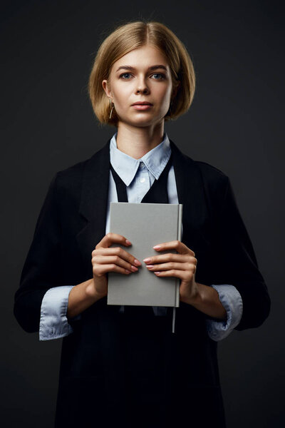 trendy blonde businesswoman holding notepad and looking at camera on black background
