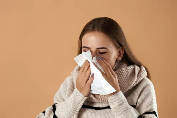 Unhappy young woman in sweater sneeze with tissue paper on beige background. suffering from flu