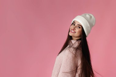 Beyaz şapkalı, pembe kazaklı güzel bir kadın pastel pembe arka plandaki kameraya bakıyor..