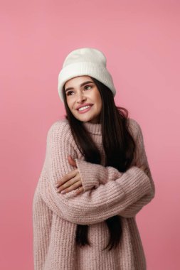 Beyaz şapkalı güzel bir kadın, kendine sarılıyor ve gülümsüyor. Pastel pembe arka planda mutlu hissediyor..