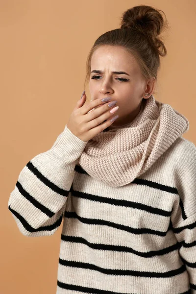 Unhappy sick woman in sweater coughing covering mouth with hand on beige background. suffering from flu