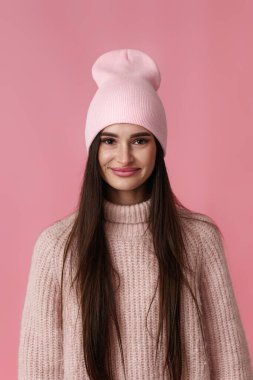 Beyaz şapkalı, pembe kazaklı genç bir kadının portresi pastel arka plandaki kameraya bakıyor..