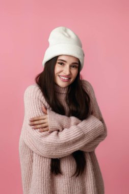 Beyaz şapkalı güzel bir kadın, kendine sarılıyor ve gülümsüyor. Pastel pembe arka planda mutlu hissediyor..