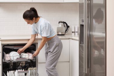 Mutlu kadın, modern beyaz mutfağın içinde temiz mutfak aletleri olan açık otomatik bulaşık makinesinin tabağını boşaltıyor..