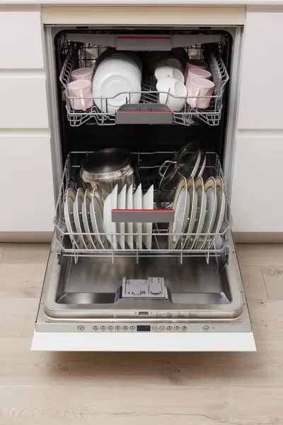 Open built-in dishwasher machine with clean cutlery, dishes, plates in white modern kitchen. front view