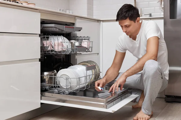 handsome man putting detergent tablet into dishwasher in kitchen