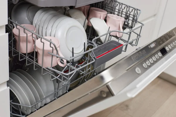Open built-in dishwasher machine with clean cutlery, dishes, plates in white modern kitchen