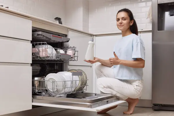 woman pours rinse aid into the dishwasher compartment in modern white kitchen