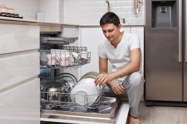 Yakışıklı, pozitif adam mutfaktaki bulaşık makinesine tabak koyuyor.