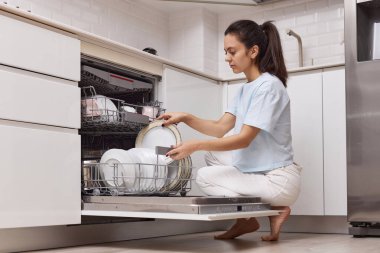 Güzel bir kadın, modern beyaz mutfağın içinde temiz mutfak aletleri olan açık otomatik bulaşık makinesinden tabakları boşaltıyor..
