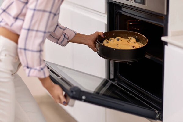женские руки кладут кастрюлю сливового теста в современную кухонную печь.