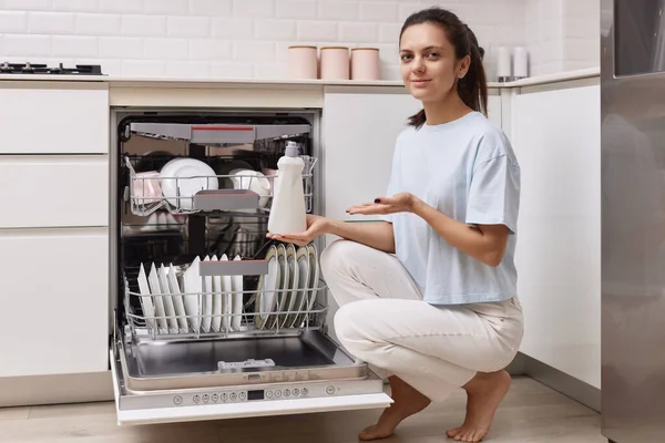 woman pours rinse aid into the dishwasher compartment in modern white kitchen