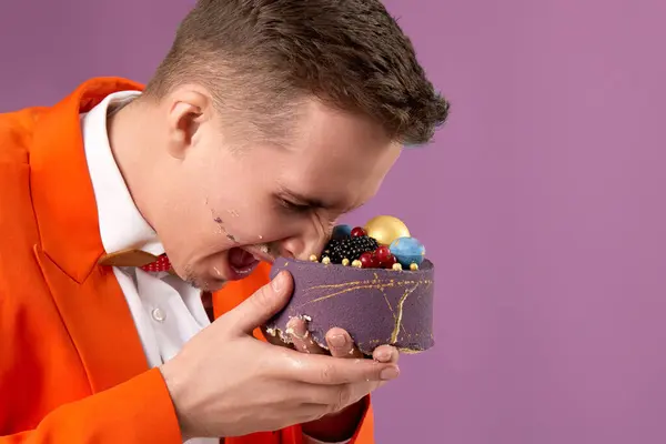 birthday man in orange jacket bites cake on purple background