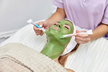 Kadın güzellik uzmanı maskesini güzellik salonundaki güzel kadının yüzüne takıyor. yüz bakımı