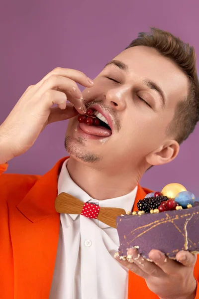 handsome birthday man in orange jacket eating tasty cake on purple background