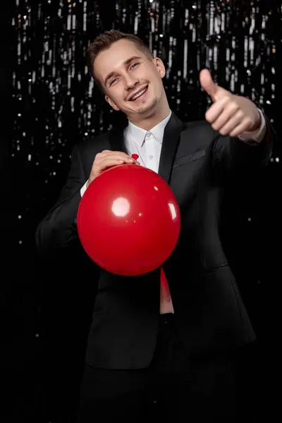 caucasian smiling man in black jacket with red air balloon showing ok gesture on dark glitter background.