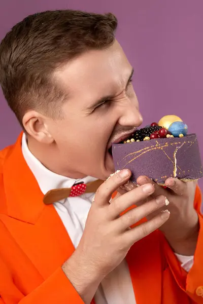 handsome birthday man in orange jacket bites cake on purple background