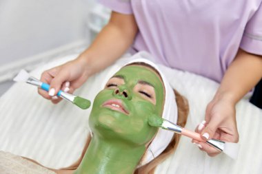 Kadın güzellik uzmanı maskesini güzellik salonundaki güzel kadının yüzüne takıyor. yüz bakımı