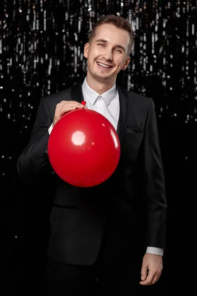 stock image smiling man in black jacket holding red air balloon on dark glitter background. new year party