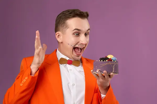 amazed birthday man in orange jacket eating cake on purple background