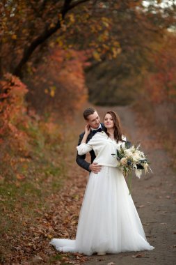 Beyaz gelinlik içinde mutlu gelin ve damat doğal arka planda dikiliyorlar.